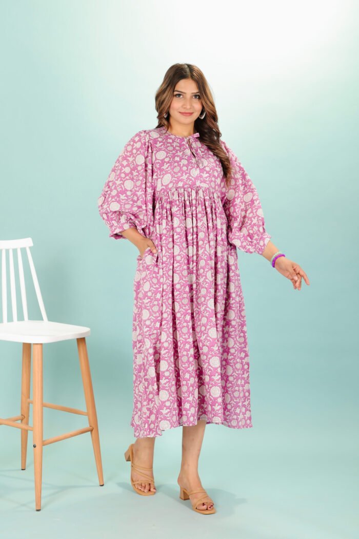 A woman in a pink floral maxi dress and with long sleeve stand in front of a light color wall