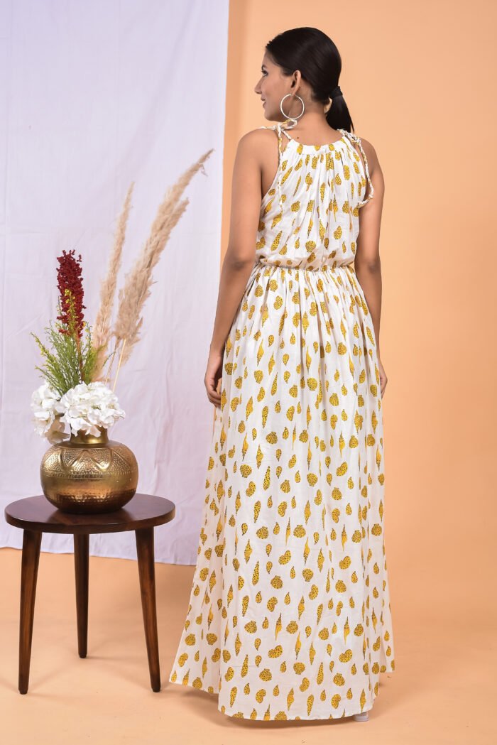 A woman in a white, floral-themed white maxi dress with a gathered waist and halter neckline stands in front of a light orange wall with a white drape and a table with a brass vase and flowers.