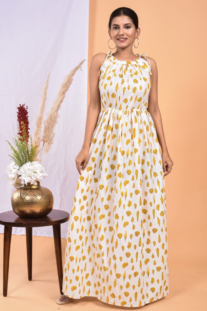 A woman in a white, floral-themed white maxi dress with a gathered waist and halter neckline stands in front of a light orange wall with a white drape and a table with a brass vase and flowers.