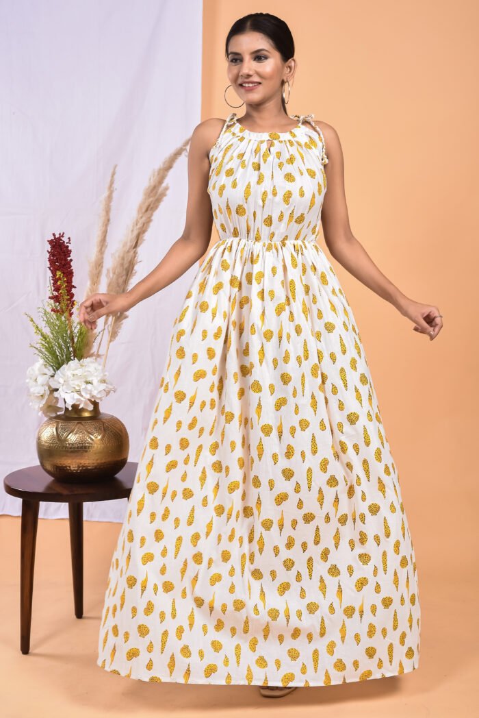 A woman in a white, floral-themed white maxi dress with a gathered waist and halter neckline stands in front of a light orange wall with a white drape and a table with a brass vase and flowers.