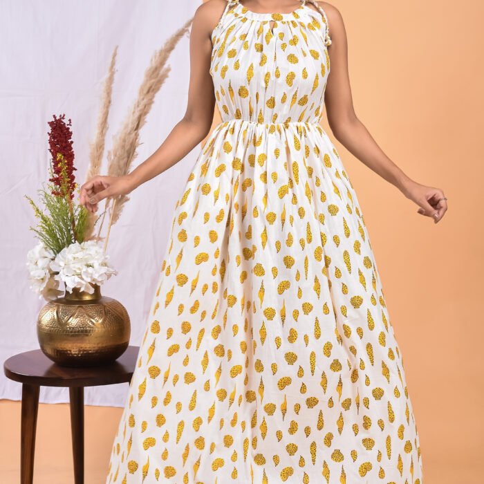 A woman in a white, floral-themed white maxi dress with a gathered waist and halter neckline stands in front of a light orange wall with a white drape and a table with a brass vase and flowers.