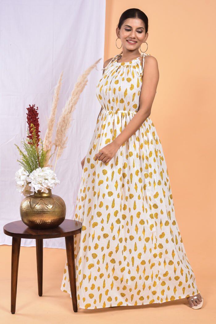 A woman in a white, floral-themed white maxi dress with a gathered waist and halter neckline stands in front of a light orange wall with a white drape and a table with a brass vase and flowers.