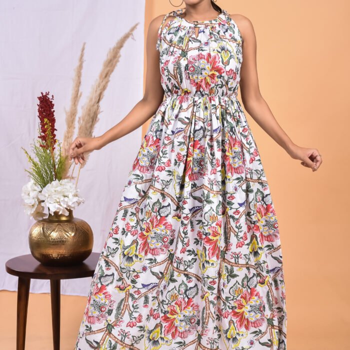 A woman in a white, floral-themed floral maxi dress with a gathered waist and halter neckline stands in front of a light orange wall with a white drape and a table with a brass vase and flowers.