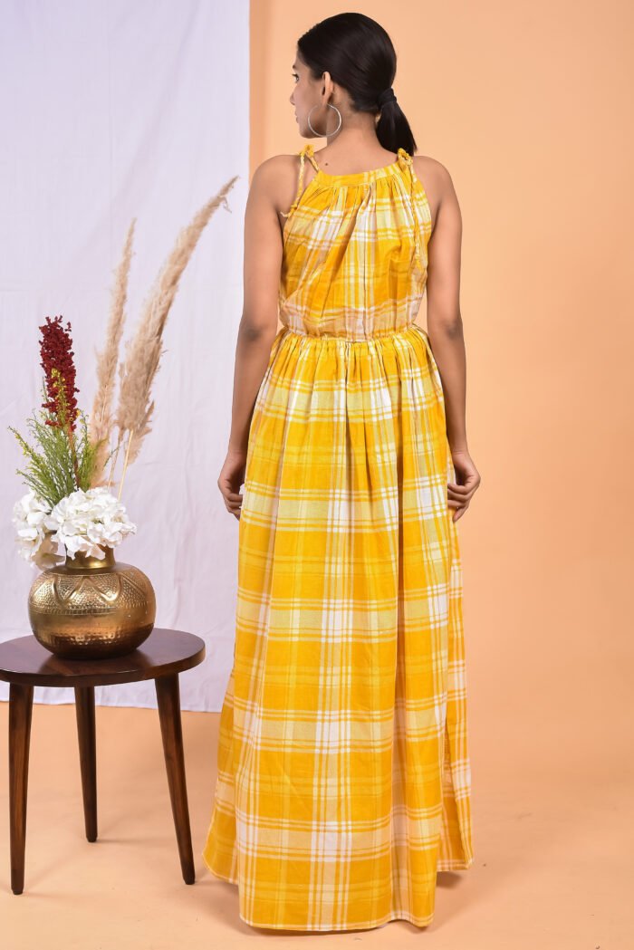 A woman in a, checks-pattern yellow maxi dress with a gathered waist and halter neckline stands in front of a light orange wall with a white drape and a table with a brass vase and flowers.