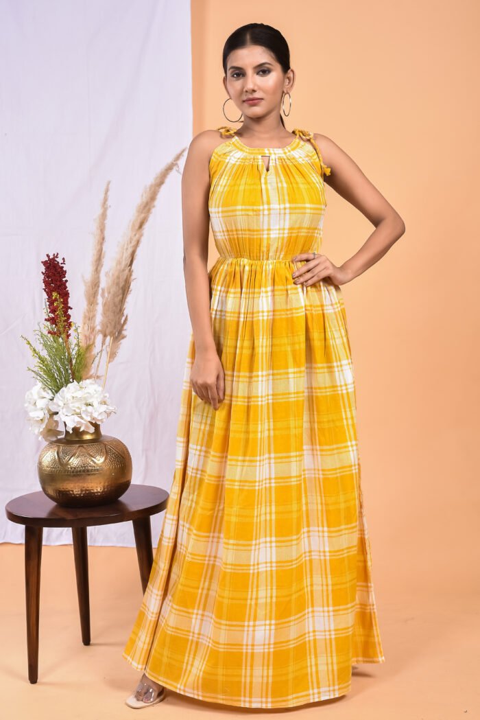 A woman in a, checks-pattern yellow maxi dress with a gathered waist and halter neckline stands in front of a light orange wall with a white drape and a table with a brass vase and flowers.