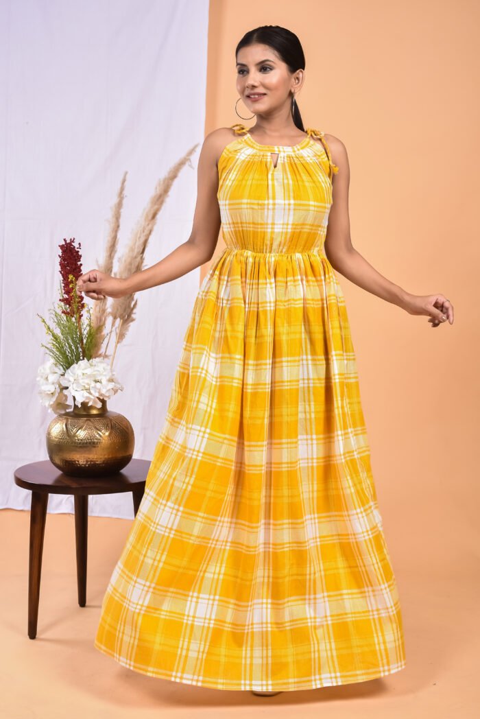 A woman in a, checks-pattern yellow maxi dress with a gathered waist and halter neckline stands in front of a light orange wall with a white drape and a table with a brass vase and flowers.