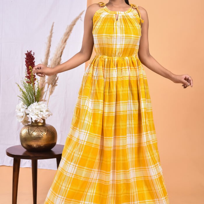 A woman in a, checks-pattern yellow maxi dress with a gathered waist and halter neckline stands in front of a light orange wall with a white drape and a table with a brass vase and flowers.