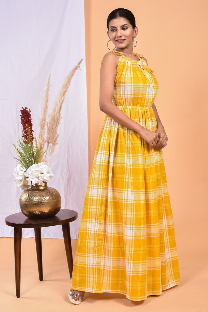 A woman in a, checks-pattern yellow maxi dress with a gathered waist and halter neckline stands in front of a light orange wall with a white drape and a table with a brass vase and flowers.