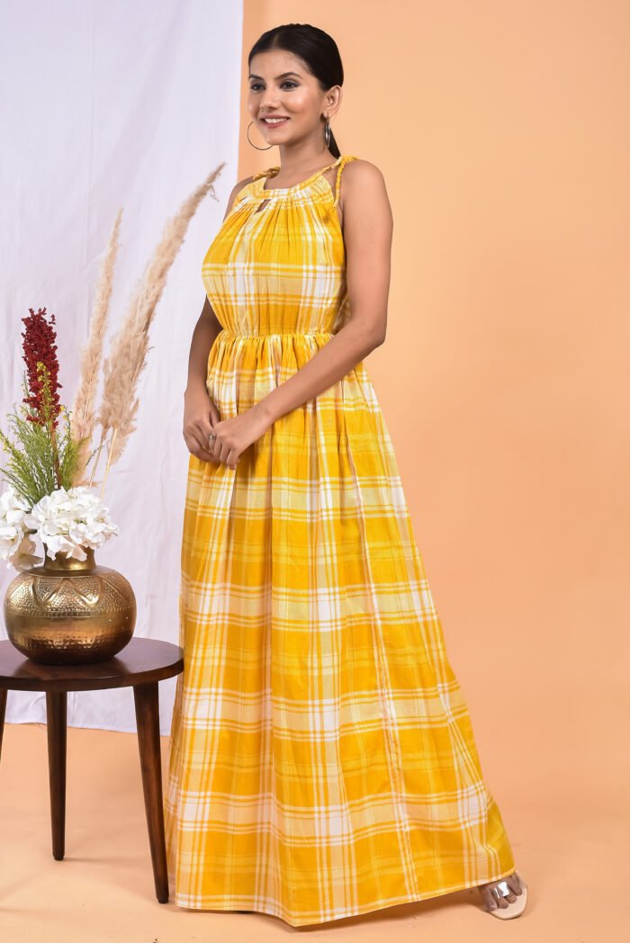 A woman in a, checks-pattern yellow maxi dress with a gathered waist and halter neckline stands in front of a light orange wall with a white drape and a table with a brass vase and flowers.