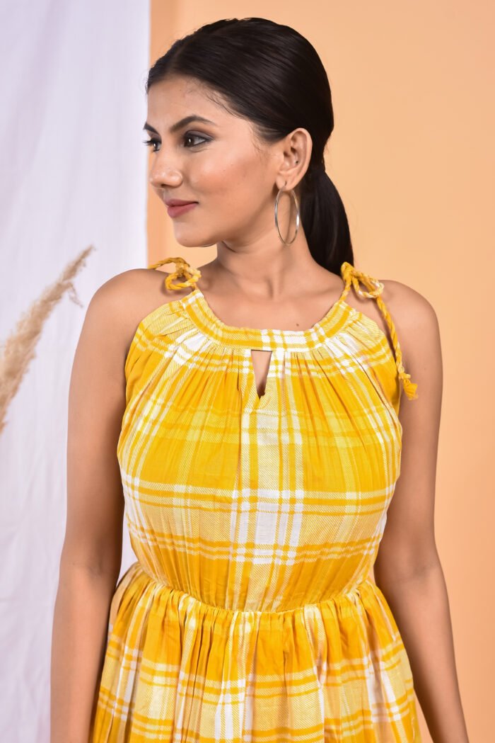 A woman in a, checks-pattern yellow maxi dress with a gathered waist and halter neckline stands in front of a light orange wall with a white drape and a table with a brass vase and flowers.