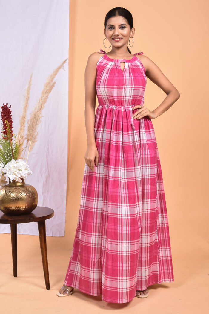 A woman in a checks-pattern pink boho maxi dress with a gathered waist and halter neckline stands in front of a light orange wall with a white drape and a table with a brass vase and flowers.