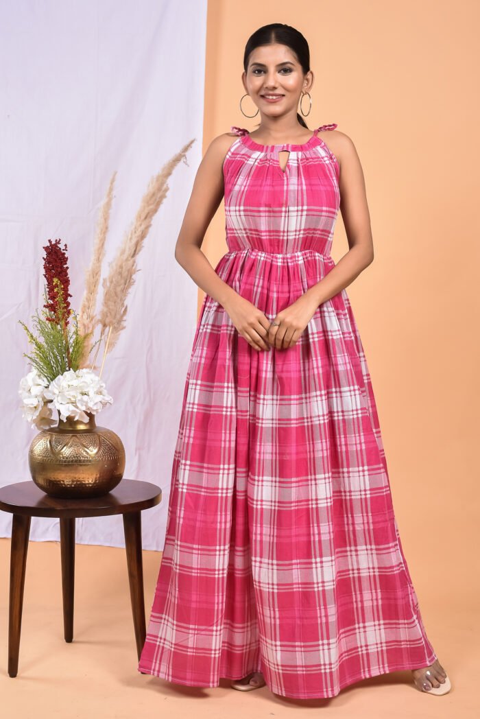 A woman in a checks-pattern pink boho maxi dress with a gathered waist and halter neckline stands in front of a light orange wall with a white drape and a table with a brass vase and flowers.