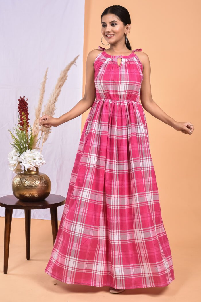 A woman in a checks-pattern pink boho maxi dress with a gathered waist and halter neckline stands in front of a light orange wall with a white drape and a table with a brass vase and flowers.