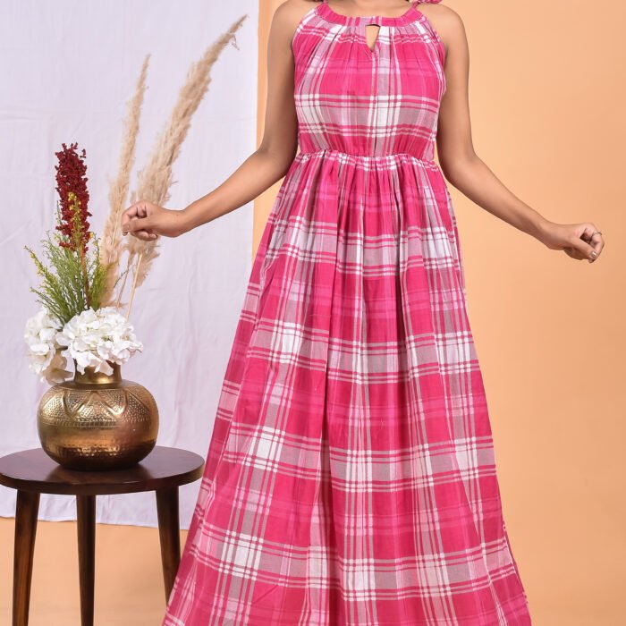 A woman in a checks-pattern pink boho maxi dress with a gathered waist and halter neckline stands in front of a light orange wall with a white drape and a table with a brass vase and flowers.