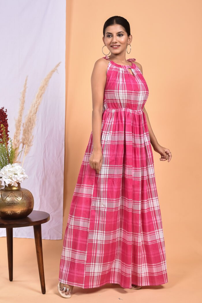 A woman in a checks-pattern pink boho maxi dress with a gathered waist and halter neckline stands in front of a light orange wall with a white drape and a table with a brass vase and flowers.