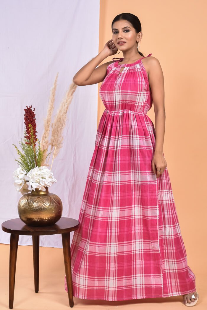 A woman in a checks-pattern pink boho maxi dress with a gathered waist and halter neckline stands in front of a light orange wall with a white drape and a table with a brass vase and flowers.