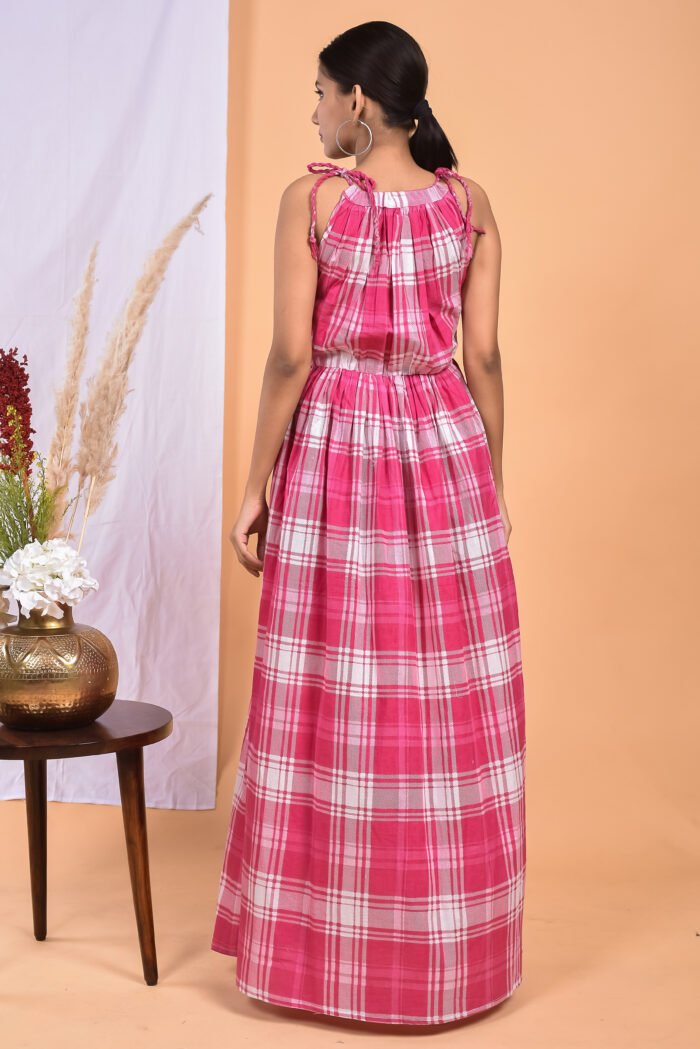 A woman in a checks-pattern pink boho maxi dress with a gathered waist and halter neckline stands in front of a light orange wall with a white drape and a table with a brass vase and flowers.