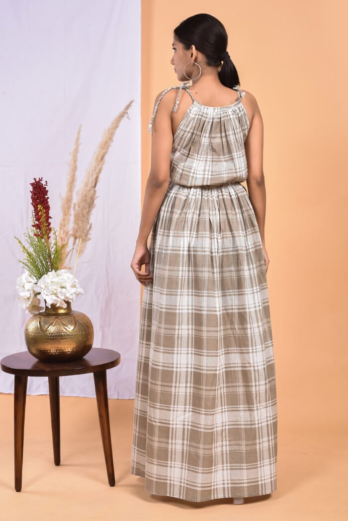 A woman in a checks pattern maxi dress with a gathered waist and halter neckline stands in front of a light orange wall with a white drape and a table with a brass vase and flowers.