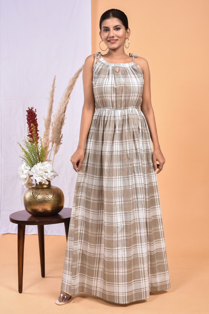 A woman in a checks pattern maxi dress with a gathered waist and halter neckline stands in front of a light orange wall with a white drape and a table with a brass vase and flowers.