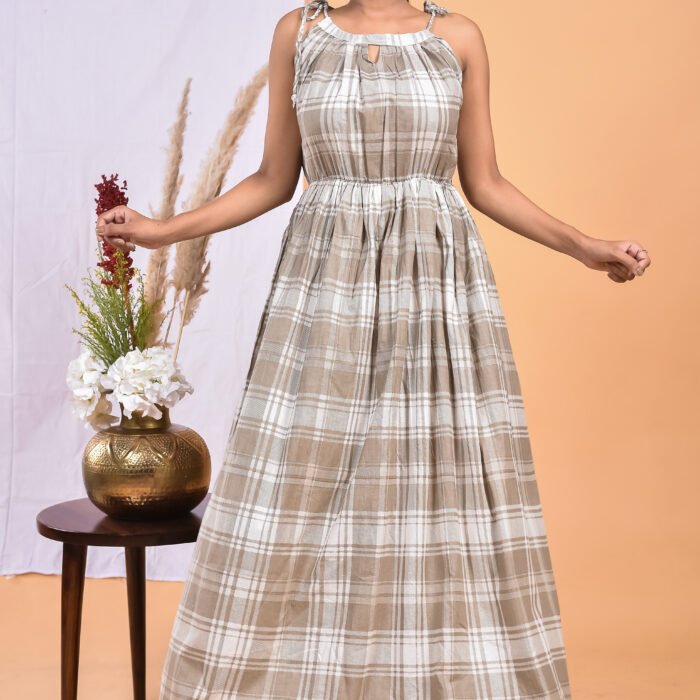 A woman in a checks pattern maxi dress with a gathered waist and halter neckline stands in front of a light orange wall with a white drape and a table with a brass vase and flowers.