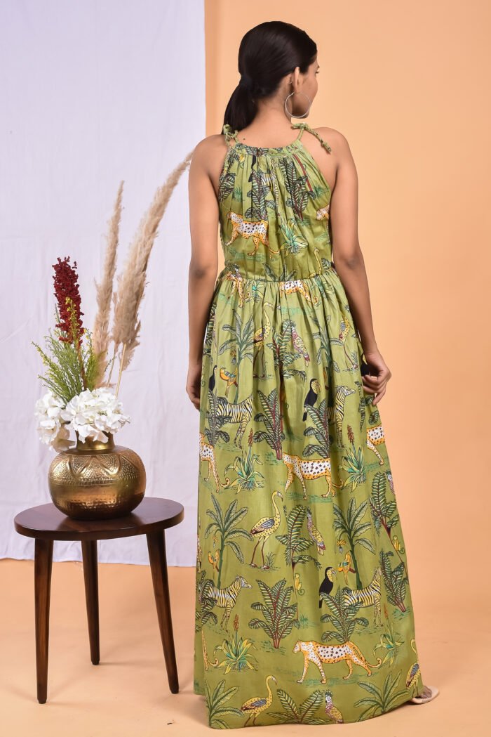 A woman in a green, jungle-themed boho maxi dress with a gathered waist and halter neckline stands in front of a light orange wall with a white drape and a table with a brass vase and flowers.