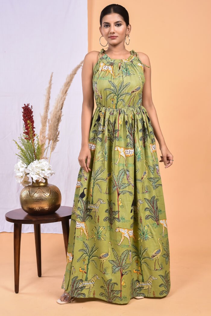 A woman in a green, jungle-themed boho maxi dress with a gathered waist and halter neckline stands in front of a light orange wall with a white drape and a table with a brass vase and flowers.