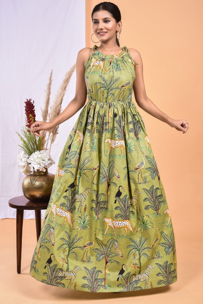 A woman in a green, jungle-themed boho maxi dress with a gathered waist and halter neckline stands in front of a light orange wall with a white drape and a table with a brass vase and flowers.