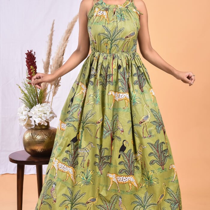 A woman in a green, jungle-themed boho maxi dress with a gathered waist and halter neckline stands in front of a light orange wall with a white drape and a table with a brass vase and flowers.