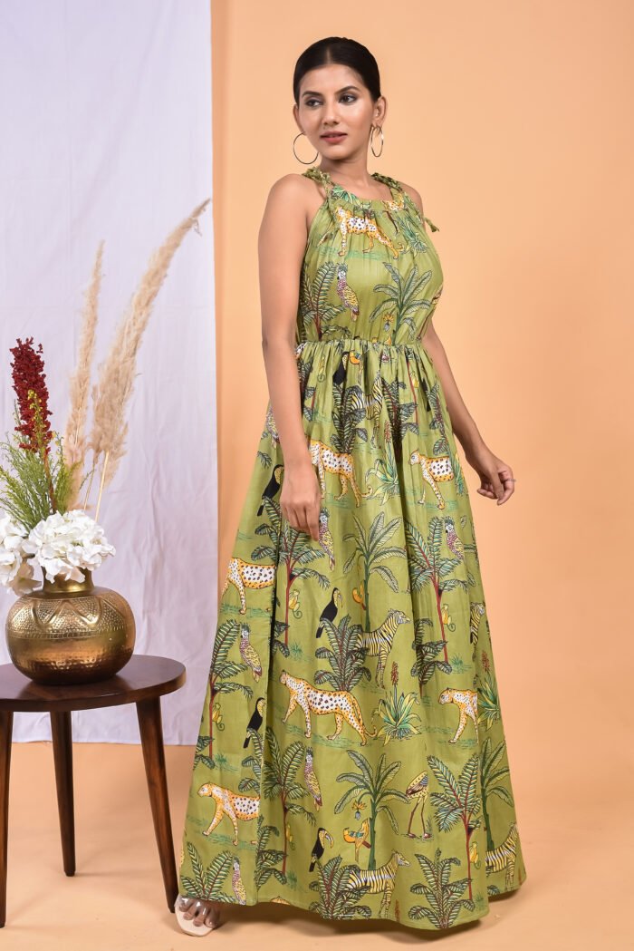 A woman in a green, jungle-themed boho maxi dress with a gathered waist and halter neckline stands in front of a light orange wall with a white drape and a table with a brass vase and flowers.