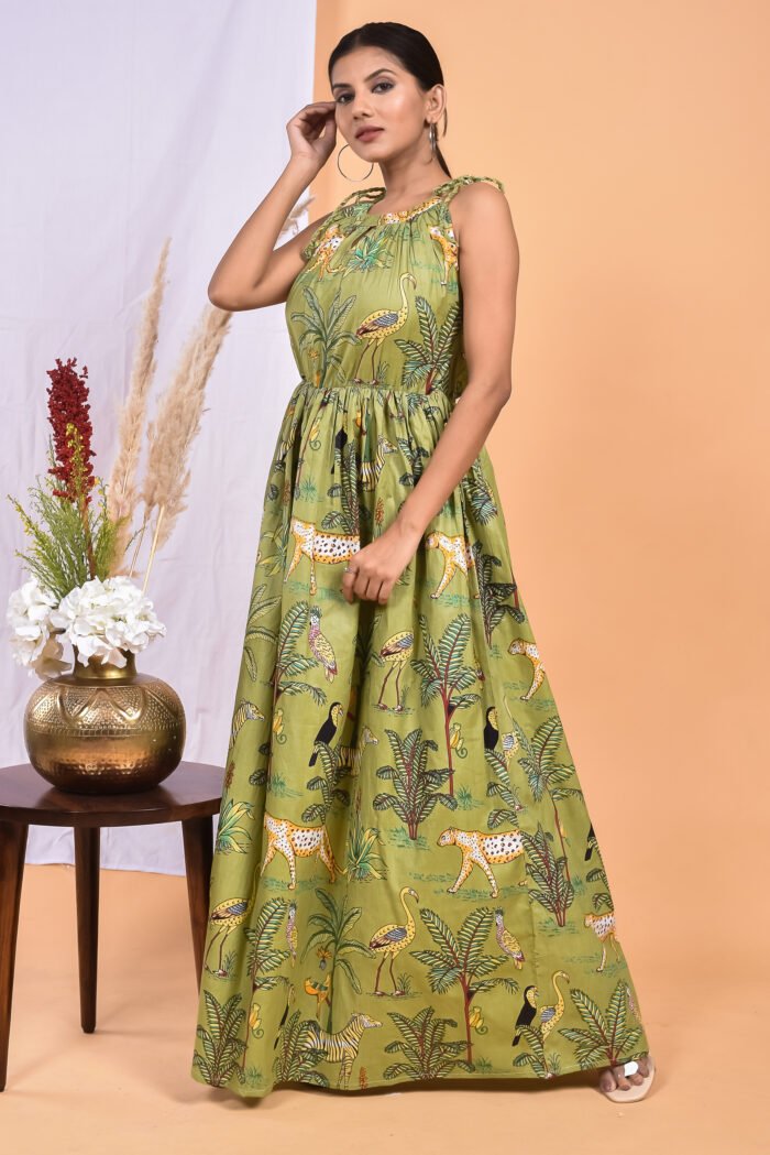 A woman in a green, jungle-themed boho maxi dress with a gathered waist and halter neckline stands in front of a light orange wall with a white drape and a table with a brass vase and flowers.