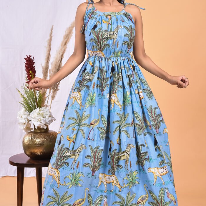 A woman in a blue maxi dress, jungle-themed with a gathered waist and halter neckline stands in front of a light orange wall with a white drape and a table with a brass vase and flowers.