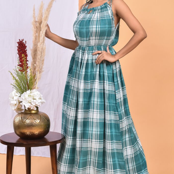 A woman in a blue & white, checks-pattern boho maxi dress with a gathered waist and halter neckline stands in front of a light orange wall with a white drape and a table with a brass vase and flowers.