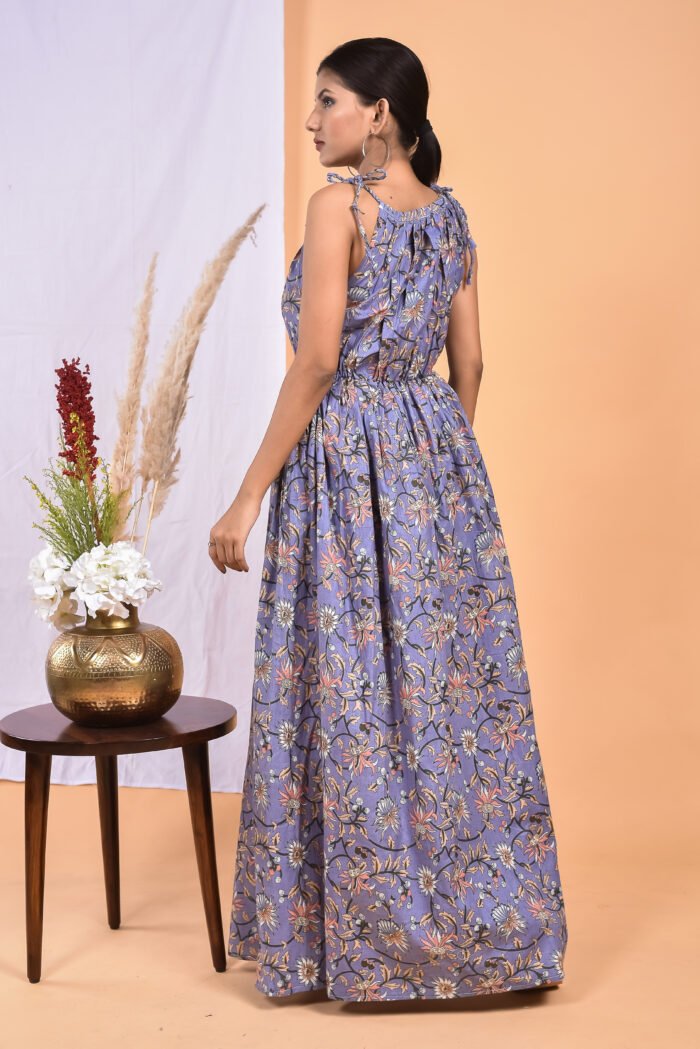 A woman in a purple, floral-themed maxi dress with a gathered waist and halter neckline stands in front of a light orange wall with a white drape and a table with a brass vase and flowers.