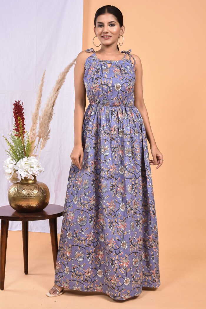 A woman in a purple, floral-themed maxi dress with a gathered waist and halter neckline stands in front of a light orange wall with a white drape and a table with a brass vase and flowers.