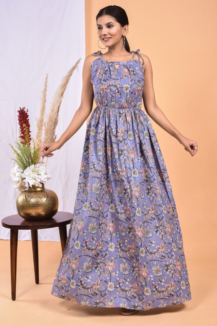 A woman in a purple, floral-themed boho maxi dress with a gathered waist and halter neckline stands in front of a light orange wall with a white drape and a table with a brass vase and flowers.