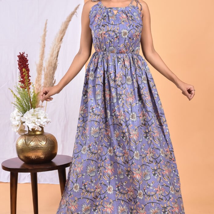 A woman in a purple, floral-themed boho maxi dress with a gathered waist and halter neckline stands in front of a light orange wall with a white drape and a table with a brass vase and flowers.