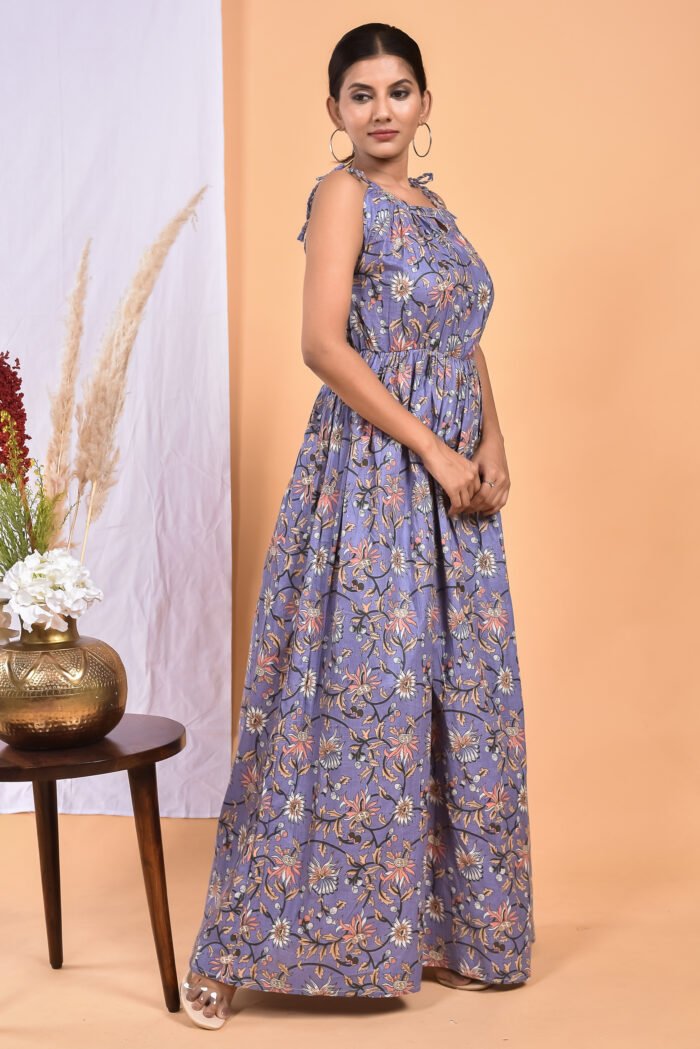 A woman in a purple, floral-themed maxi dress with a gathered waist and halter neckline stands in front of a light orange wall with a white drape and a table with a brass vase and flowers.