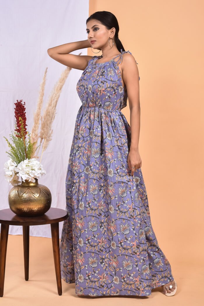 A woman in a purple, floral-themed maxi dress with a gathered waist and halter neckline stands in front of a light orange wall with a white drape and a table with a brass vase and flowers.