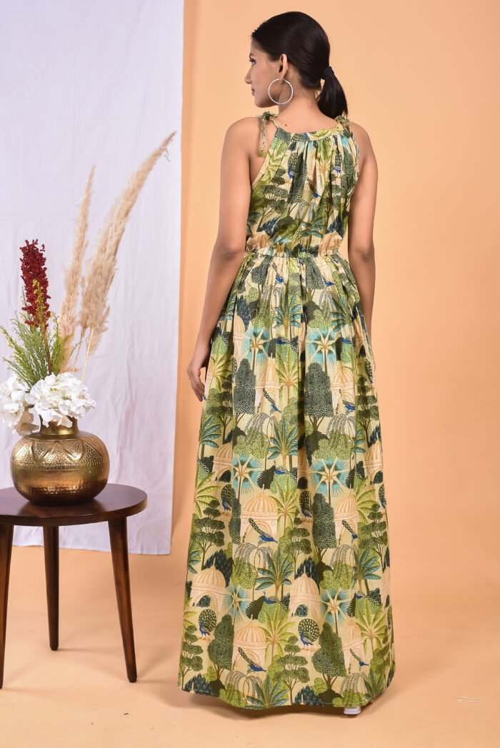 A woman in a green, jungle-themed maxi dress with a gathered waist and halter neckline stands in front of a light orange wall with a white drape and a table with a brass vase and flowers.