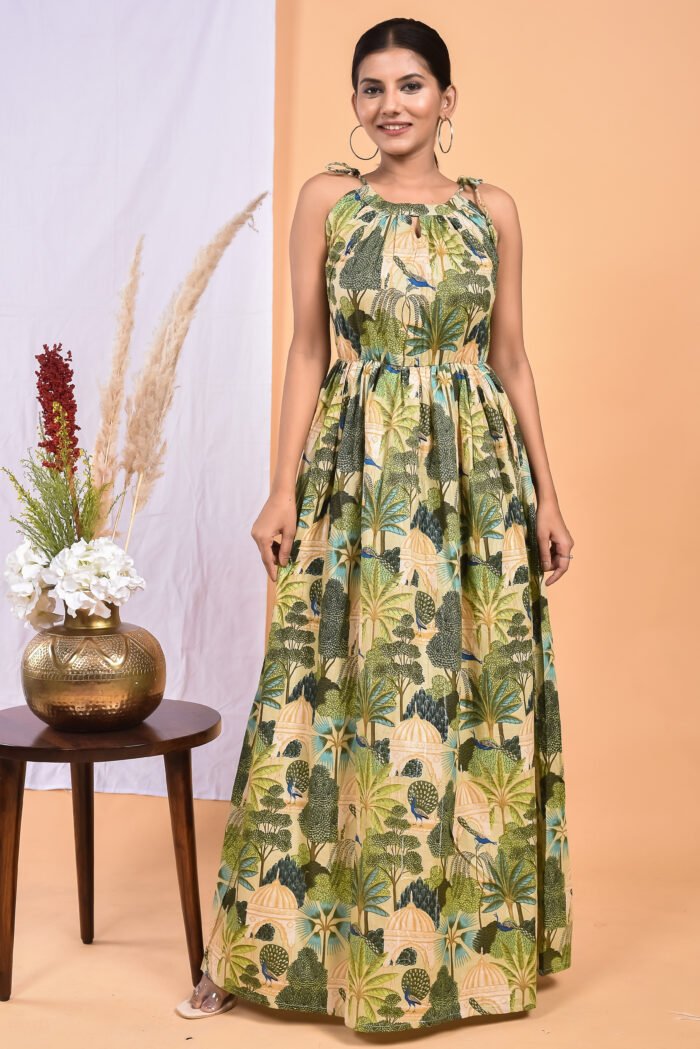 A woman in a green, jungle-themed maxi dress with a gathered waist and halter neckline stands in front of a light orange wall with a white drape and a table with a brass vase and flowers.