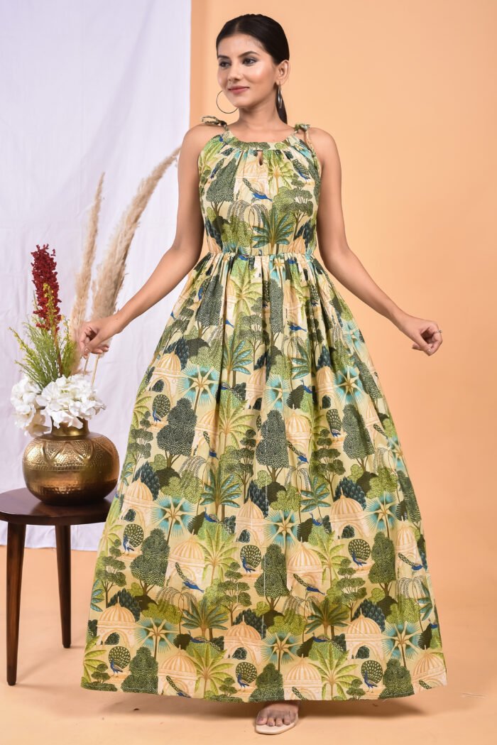 A woman in a green, jungle-themed boho maxi dress with a gathered waist and halter neckline stands in front of a light orange wall with a white drape and a table with a brass vase and flowers.