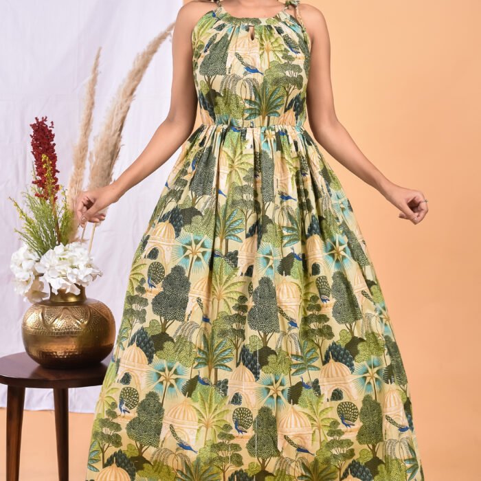 A woman in a green, jungle-themed boho maxi dress with a gathered waist and halter neckline stands in front of a light orange wall with a white drape and a table with a brass vase and flowers.