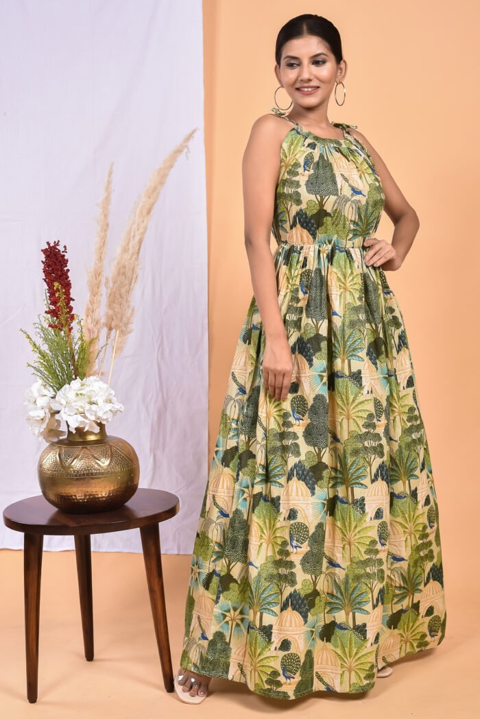 A woman in a green, jungle-themed maxi dress with a gathered waist and halter neckline stands in front of a light orange wall with a white drape and a table with a brass vase and flowers.