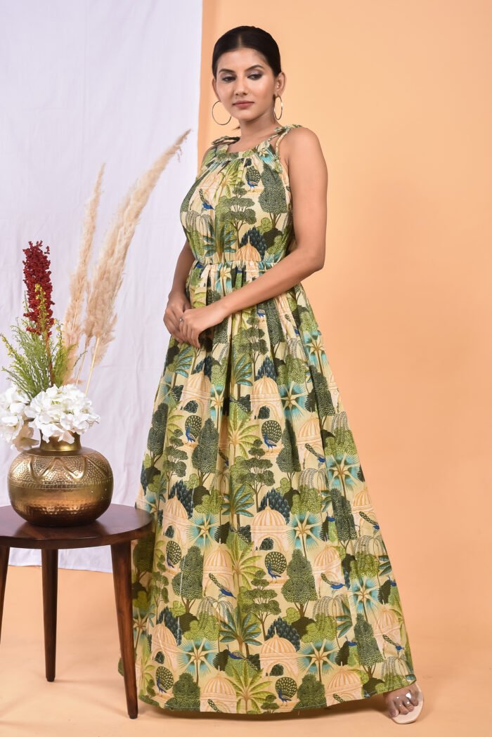 A woman in a green, jungle-themed maxi dress with a gathered waist and halter neckline stands in front of a light orange wall with a white drape and a table with a brass vase and flowers.